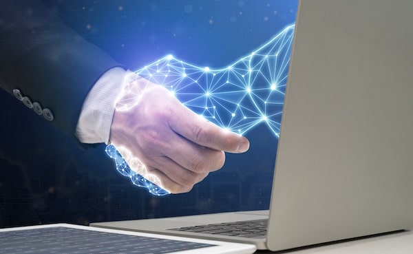 Digital handshake: A glowing wireframe hand emerges from a laptop screen, shaking hands with a human hand, symbolizing AI-driven connection and co-intelligence.