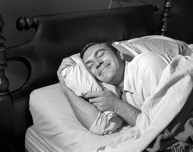 A man in pajamas lies in bed, smiling contentedly while hugging a pillow with both arms, showing how simple habits can help you improve your sleep.