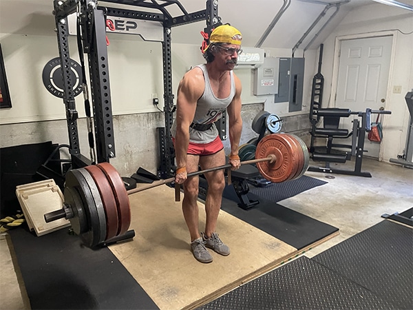 A person wearing a yellow bandana and glasses is performing a deadlift with a loaded barbell in a home gym, showcasing their dedication to fitness and exercise.