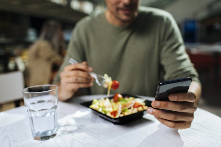 A person sits at a table eating a salad and using a smartphone, perhaps messaging their personal nutrition coach for tips, with a glass of water nearby. The focus is on their hands and the plate of food.
