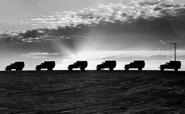 Silhouetted trucks drive in line along a ridge at sunset, demonstrating precise convoy execution against a backdrop of a cloudy sky and beams of sunlight.