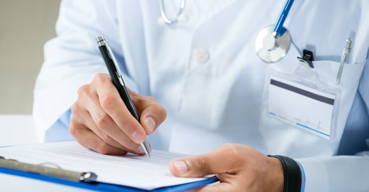 A doctor wearing a lab coat and stethoscope writes on a clipboard form, with a blank name badge visible.
