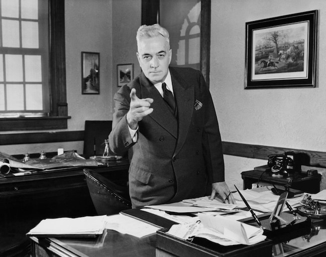 A man in a suit stands behind a desk covered with papers and points directly at the camera, embodying the confidence needed to start executing plans in an office setting.