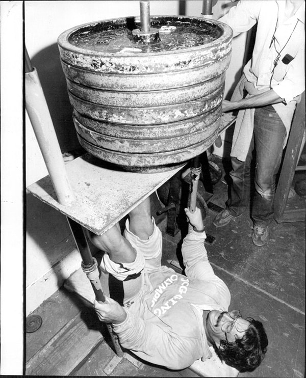 A man lies on his back performing a classic Mike Mentzer workout, holding a platform loaded with heavy weights and lifting it upwards as another person stands nearby assisting.
