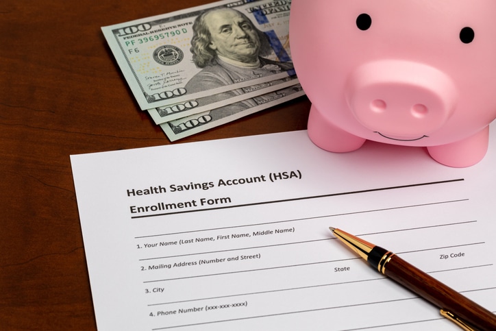 A Health Savings Account enrollment form sits on a desk next to a pen, three $100 bills, and a pink piggy bank—ready to help you build wealth for your six-figure future.