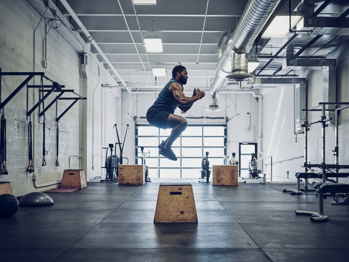 A man showcases his explosive strength with a box jump in the gym, where he's surrounded by weights, exercise balls, and other fitness equipment. Clad in athletic gear, he defies powerpenia with every leap.