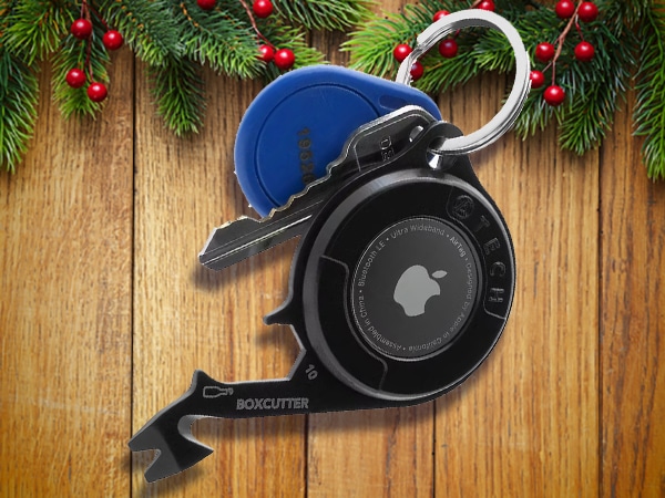 A keychain with keys, a blue fob, a black Apple AirTag, and a multi-tool rests on wooden boards with pine and red berries in the background—perfect stocking stuffers for men this holiday season.